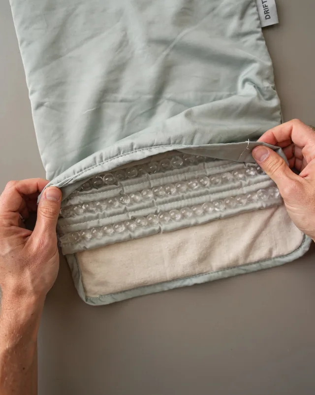 A pair of hands peels back the corner of a folded sage-green blanket tagged ‘Drift’ to reveal its internal layers: a row of glass beads sewn into channels over a plain base fabric.