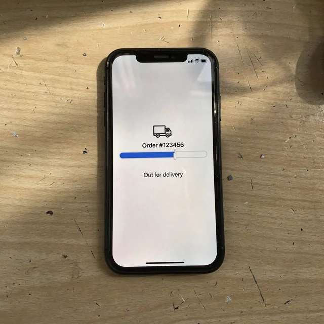 A phone lying flat on a wood desk, its screen showing a plain order-tracking mock: a truck icon, ‘Order #123456,’ a progress bar and ‘Out for delivery.’